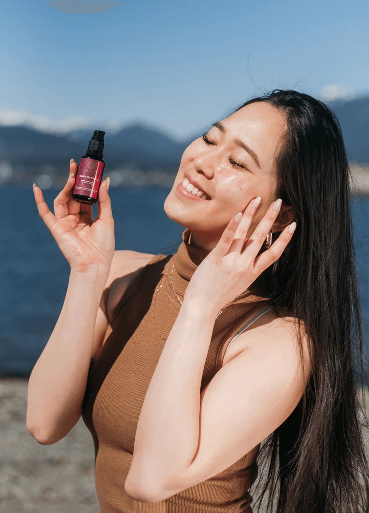 Smiling woman outdoors holding OKOKO L’Élixir de Pureté Dragon’s Blood and Niacinamide serum, enjoying radiant skin care under natural light in Vancouver